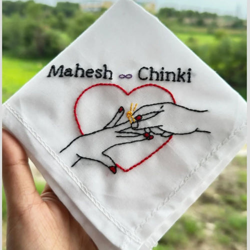 Embroidered fabric with hands holding a ring and a heart, with 'customized couple names' embroidered above, held against a blurred green outdoor background.